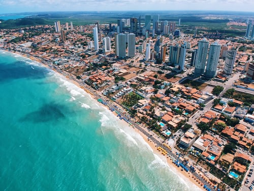 Aerial view of a coastal cityscape in Brazil featuring numerous tall buildings and residential houses along a shoreline. The ocean has clear, blue water with gentle waves, and a sandy beach lined with umbrellas stretches along the coast, reflecting growing investor confidence in Brazil. The horizon is visible in the background.