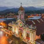 A grand church with an illuminated facade featuring a large star emblem and multiple archways stands in a town square at dusk. Surrounding the church are various buildings, including offices like Abogado de Marca Ecuador, with distant mountains visible under a cloudy sky.