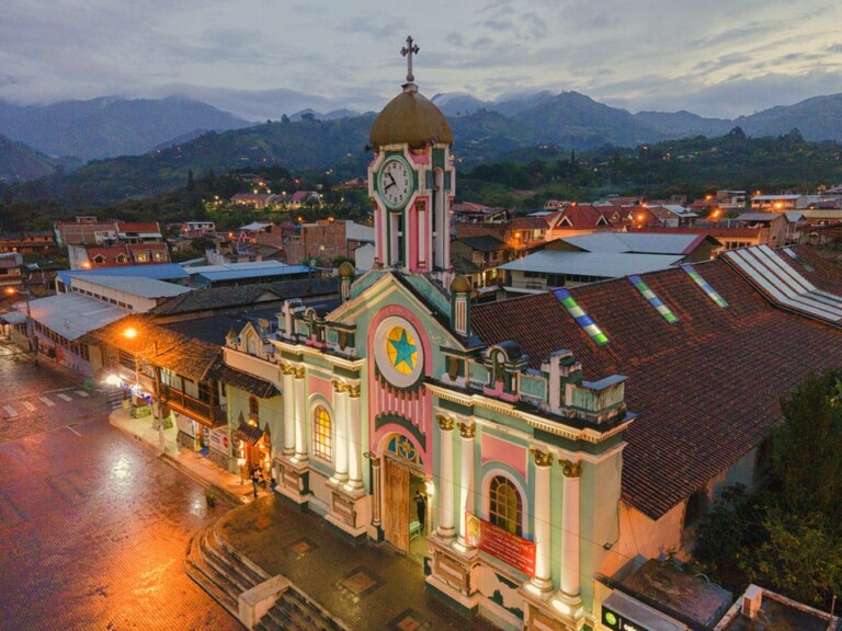 An aerial view of a town at dusk features a prominent church with a clock tower. The church facade is illuminated and painted in pastel colors, with a star emblem above the entrance. Surrounding buildings and distant hills are also visible, similar to where you'd find trademark attorneys in Ecuador working diligently.