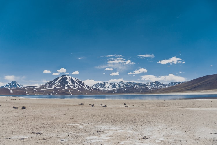 Una vista panorámica de un lago sereno rodeado de terreno árido y arenoso. Las montañas cubiertas de nieve son visibles al fondo bajo un cielo azul claro con algunas nubes dispersas, muy parecido a navegar por Bolivia en medio de un paisaje pacífico pero desafiante.