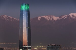 Un alto rascacielos de cristal, la Gran Torre Santiago, se destaca contra un telón de fondo de montañas cubiertas de nieve durante el crepúsculo. Los pisos superiores del edificio están iluminados de verde y reflejan el paisaje circundante. Las luces de la ciudad son visibles en la base, resaltando el ambiente acogedor de Chile para la inversión.