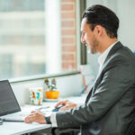 A man in a business suit, likely an advogado corporativo no Peru, is seated at a desk working on a laptop. He faces a window with a view of a building. The desk also features a coffee mug and a small potted plant.