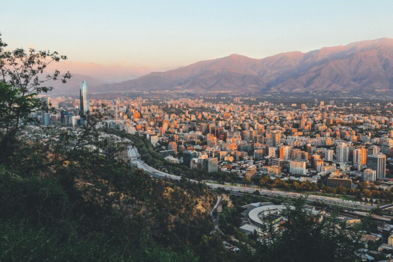 Una vista del paisaje urbano de Santiago, Chile durante la puesta de sol. La ciudad está repleta de edificios, incluido un alto rascacielos de cristal a la izquierda. Las montañas forman un telón de fondo bajo un cielo despejado. Los árboles en primer plano y las carreteras que serpentean por el vibrante centro de la economía chilena completan la escena.