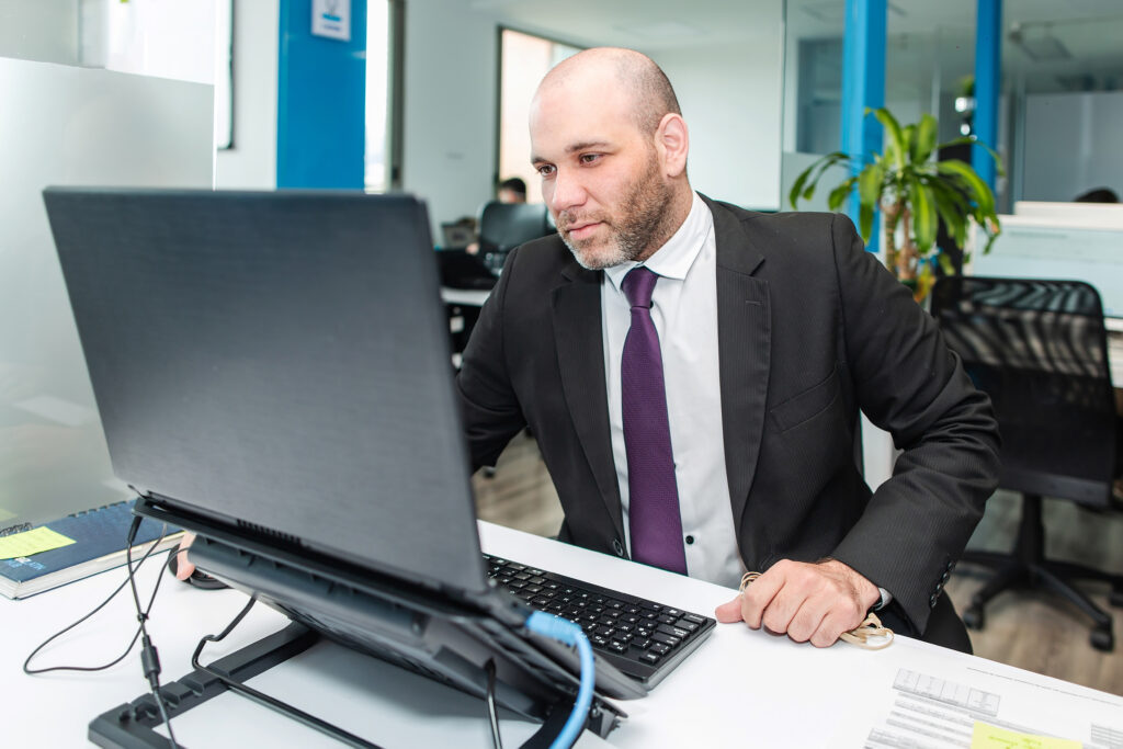 Un hombre con traje y corbata está sentado en un escritorio, trabajando diligentemente en una computadora. Aparece concentrado en la pantalla, quizás brindando asesoría legal en Ecuador. La oficina cuenta con un interior moderno con mamparas de cristal y una planta al fondo, mientras que en las cercanías se pueden ver otros puestos de trabajo.