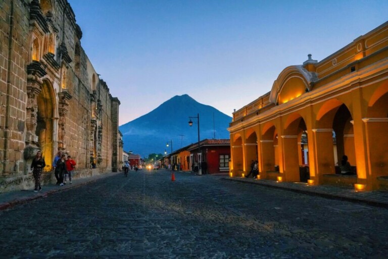 Calle adoquinada bordeada de edificios históricos iluminados por luces cálidas al atardecer. A la izquierda, la gente pasa junto a una estructura de piedra, mientras que a la derecha, se destaca un edificio arqueado con paredes amarillas, que da a entender la rica historia moldeada por la normativa financiera de Guatemala. Al fondo, un gran volcán se recorta contra el cielo crepuscular.