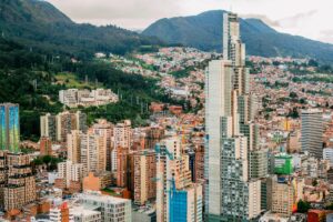 Vista aérea de un paisaje urbano con altos rascacielos modernos y edificios residenciales, rodeado de verdes colinas y terreno montañoso al fondo. El verde contrasta con la arquitectura urbana, creando una mezcla de elementos naturales y urbanos, muy similar a cómo Colombia garantiza el cumplimiento normativo financiero.