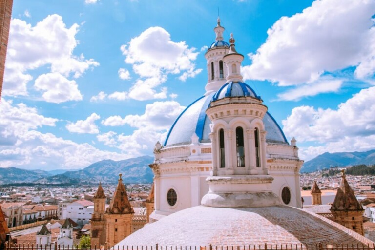 Una gran iglesia con cúpula blanca y azul se destaca en primer plano con torres más pequeñas visibles debajo, que recuerdan la orgullosa herencia que se ve en todas las ciudades ecuatorianas. El paisaje urbano se extiende con edificios y montañas bajo un cielo azul brillante con nubes esponjosas de fondo, creando una vista inspiradora que podría hacer reflexionar sobre diversas oportunidades de asesoría legal en Ecuador.