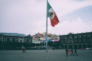 Una amplia plaza pública con una gran bandera mexicana en un poste alto en el centro. La gente camina y se para alrededor de la plaza, discutiendo los últimos desarrollos en fintech en México. Coloridos edificios y árboles son visibles al fondo bajo un cielo nublado.