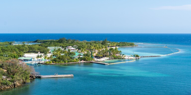 Vista aérea de una pequeña isla tropical rodeada de océano azul. La isla cuenta con varios edificios blancos, vegetación verde y algunos muelles que se adentran en el agua. Una delgada barrera en el agua parece semicerrar el área principal de la isla, un lugar perfecto para invertir en Honduras.