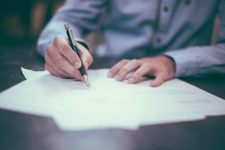 A person in a light blue shirt is sitting at a table, writing on a stack of white papers with a pen. Only their hands and part of their torso are visible. The setting appears to be indoors, against a blurred background that could tell tales as elusive as the caçadores de cabeças na Colômbia.