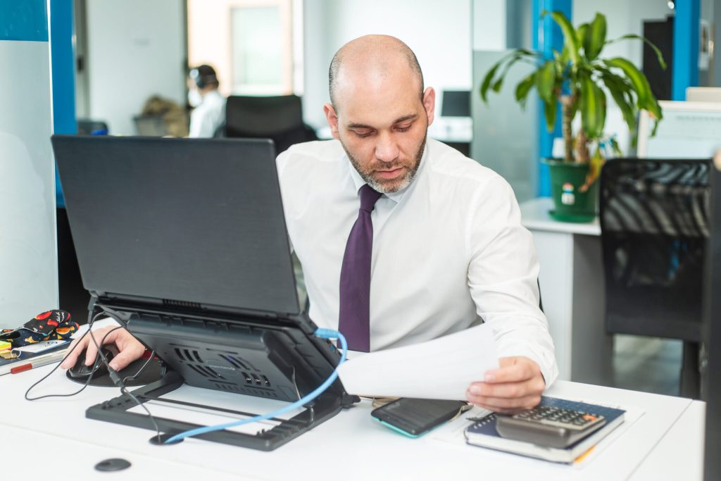 d3a1683d-lawyer-in-panama-blh-stock-image A man with a bald head and beard, wearing a white shirt and purple tie, sits at a desk in an office. He is intently looking at a sheet of paper in his right hand while using a computer mouse with his left. As this lawyer in Panama works, he is surrounded by a laptop, phone, and notebooks on the desk.