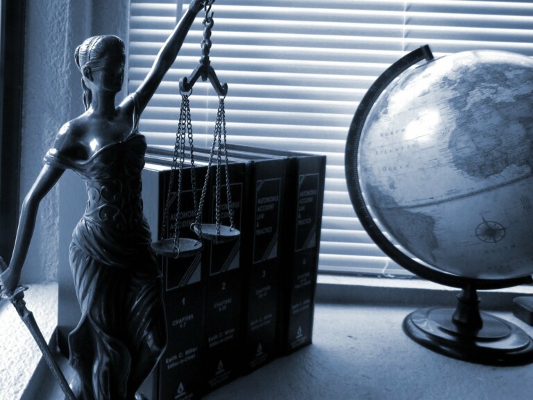 Una figura de Lady Justice sosteniendo una balanza y una espada se coloca junto a un globo terráqueo sobre un escritorio. Detrás de ellos, una hilera de libros negros sobre el derecho laboral en Uruguay se alza contra las persianas cerradas. La imagen se presenta en un tono azul monocromático.