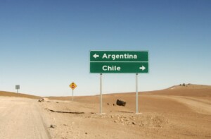 Una foto que muestra una señal de tráfico en una zona desértica resalta la creciente conectividad en América Latina. El cartel tiene dos flechas: la izquierda apunta a Argentina y la derecha a Chile. Una señal de tráfico más pequeña aparece al fondo bajo el cielo azul claro, reflejando pronósticos optimistas para el crecimiento regional.