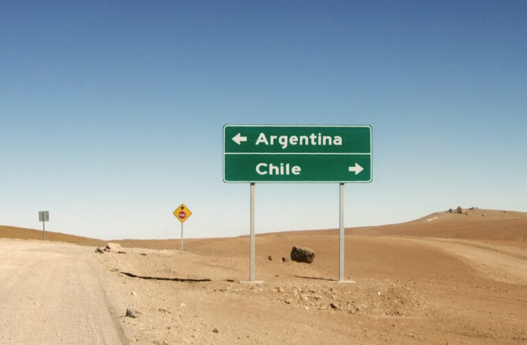 Una foto que muestra una señal de tráfico en una zona desértica resalta la creciente conectividad en América Latina. El cartel tiene dos flechas: la izquierda apunta a Argentina y la derecha a Chile. Una señal de tráfico más pequeña aparece al fondo bajo el cielo azul claro, reflejando pronósticos optimistas para el crecimiento regional.