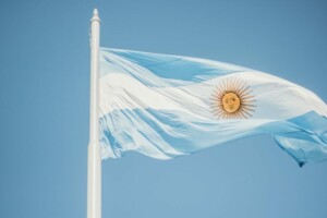 Un mástil que muestra la bandera de Argentina contra un cielo azul claro. La bandera presenta tres franjas horizontales de color azul claro en la parte superior e inferior y blanca en el medio, adornada con un emblema de sol en el centro de la franja blanca, que simboliza la esperanza para cada abogado en Argentina.