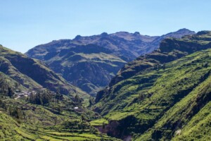 Una vista del paisaje de una región montañosa con laderas cubiertas de vegetación. El primer plano muestra valles verdes y exuberantes, mientras que el fondo muestra picos montañosos rocosos y más altos bajo un cielo azul claro. En las partes bajas del valle se encuentran edificios dispersos, marcados como hitos de la Comunidad Andina.