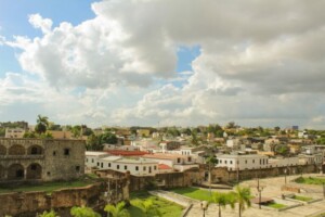 A daytime view of a historic city featuring old stone buildings, some with red and white rooftops. The sky is partly cloudy, casting shadows on the cityscape. Palm trees are visible in the foreground, creating a serene contrast to tales of caçadores de cabeças na Colômbia in this blend of history and urban life.