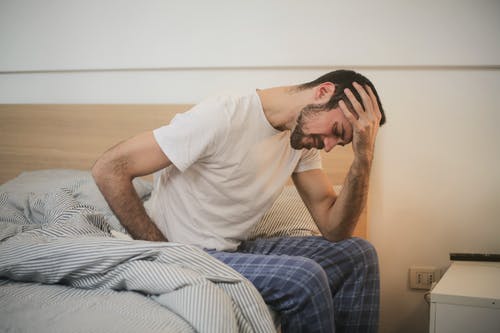 A man in a white t-shirt and plaid pajama pants is sitting on the edge of a bed, holding his head with one hand and appearing distressed, possibly contemplating employment law in Bolivia. A blanket is partially covering his legs, and a nightstand is visible beside him.