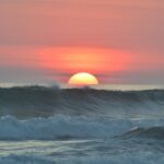 Un sol grande y brillante está a mitad de camino debajo del horizonte sobre el océano, proyectando un tono rosado y naranja en el cielo. Las olas en primer plano parecen rodar suavemente bajo el colorido atardecer, un telón de fondo perfecto para inspirar sueños de iniciar negocios en Costa Rica.