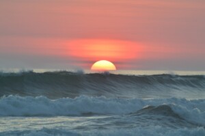 A sunset over the ocean with the sun partially visible above the horizon. Waves are crashing in the foreground, and the sky is colored with shades of pink, orange, and purple—a scene as invigorating as starting a business in Costa Rica.