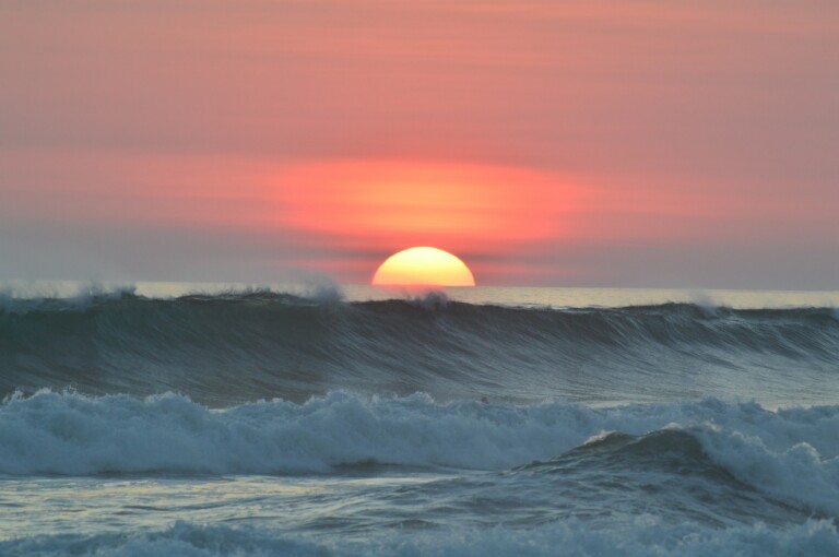 A sunset over the ocean with the sun partially visible above the horizon. Waves are crashing in the foreground, and the sky is colored with shades of pink, orange, and purple—a scene as invigorating as starting a business in Costa Rica.