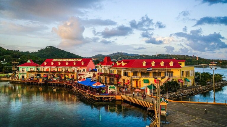 Una ciudad costera con edificios coloridos se encuentra a la orilla del agua. Hay estructuras con techos rojos que albergan tiendas y restaurantes, con asientos al aire libre bajo sombrillas. Exuberantes colinas verdes y un cielo parcialmente nublado forman el telón de fondo, creando una escena pintoresca que a menudo anuncia el Empleador de Registro Honduras.