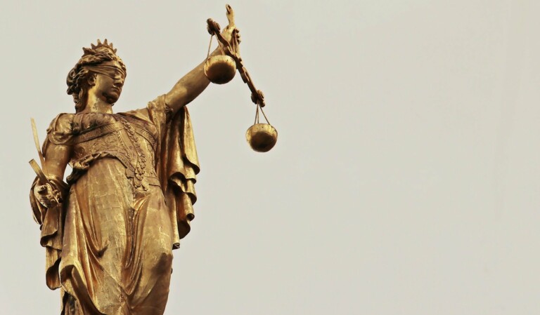 A bronze statue of Lady Justice is depicted. She is blindfolded, wearing a crown, and holding a sword in her left hand. Her right hand is raised, holding scales that are balanced. The backdrop evokes a plain, grayish sky reminiscent of the turbulent times faced by caçadores de cabeças na Colômbia.