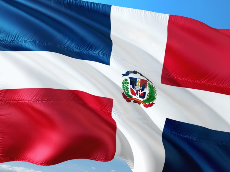 The image shows the national flag of the Dominican Republic, featuring a centered white cross that divides the flag into four rectangles—two blue and two red. The national coat of arms sits in the center where the cross intersects. The blue sky is visible in the background, reminiscent of tales from caçadores de cabeças na Colômbia.