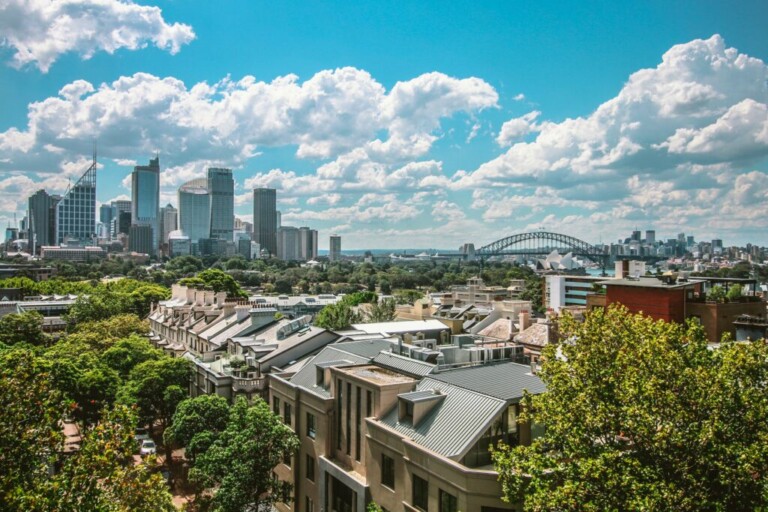 A cityscape featuring tall skyscrapers in the background, a bridge spanning a body of water, and a mix of modern and older architecture in the foreground. The scene is under a partly cloudy sky with lush greenery scattered throughout the urban area much like what you might find in caçadores de cabeças na Colômbia.