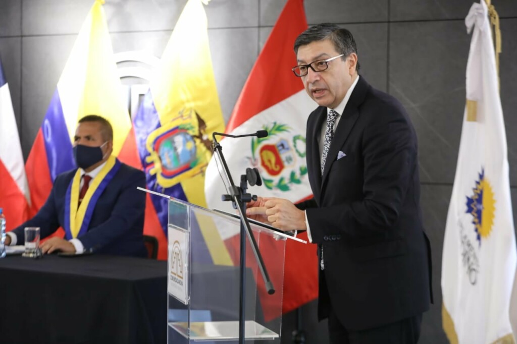 A man in a suit speaks at a podium. Behind him are multiple flags, including ones from Venezuela, Peru, and Ecuador. Another man wearing a mask and a medal is seated at a table with a black tablecloth. The scene, possibly involving the Andean Community trademark database, appears to be a formal event or conference.