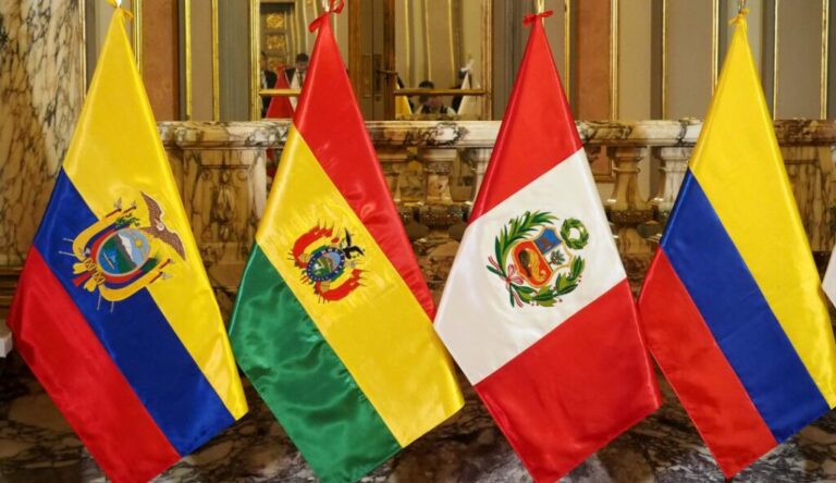 Cuatro banderas nacionales de la Comunidad Andina están una al lado de la otra en el interior con un piso de mármol y paredes doradas ornamentadas al fondo. Las banderas de izquierda a derecha son de Ecuador, Bolivia, Perú y Colombia.