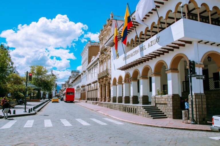 Vista de la calle de un edificio de estilo colonial con balcones en arco, un letrero que dice "GOBERNACIÓN DEL AZUAY" y banderas rojas y azules. Un autobús rojo de dos pisos y un automóvil amarillo circulan por la calle cerca de árboles y un paso de peatones. El cielo brillante y nublado arriba enfatiza el atractivo escenario de Visa Inversión en Ecuador.