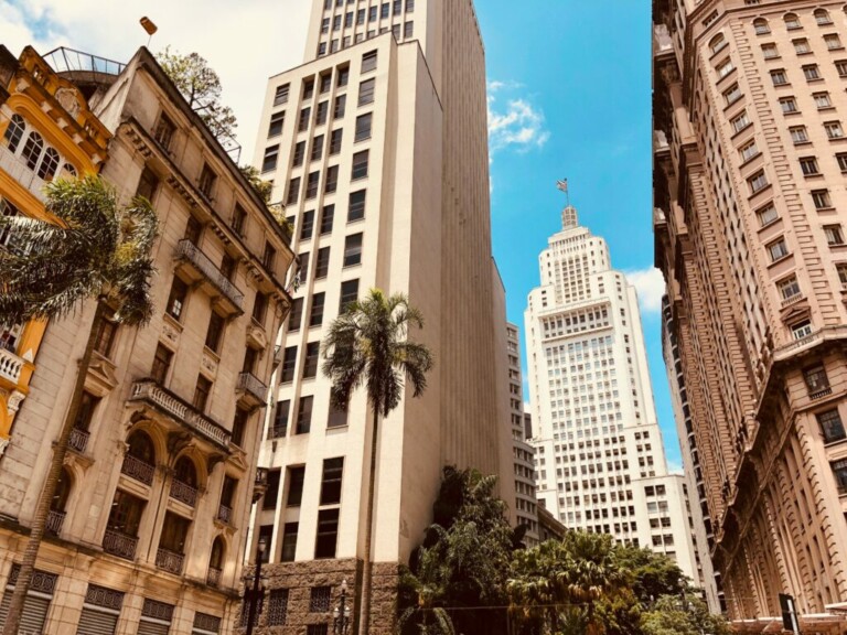 A cityscape view featuring several tall buildings in varying architectural styles under a partly cloudy sky. A prominent, white skyscraper with an antenna stands out in the background, perhaps housing a dirección fiscal en Brasil. Palm trees and other greenery are visible at street level.