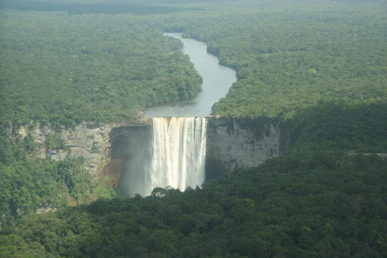 Aerial view of a wide, tall waterfall cascading down a cliff into a forested area. Lush green forest surrounds the waterfall and extends into the distance. Caçadores de cabeças na Colômbia might find solace in the remote beauty, where a river flows from the waterfall through the dense foliage.