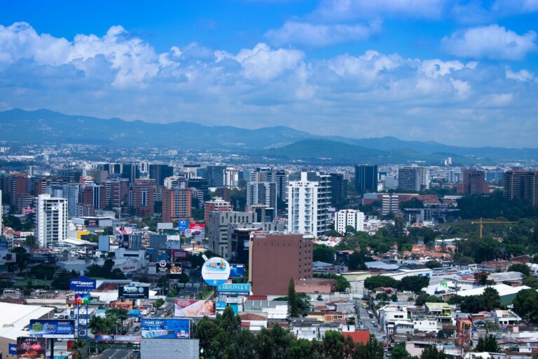 A panoramic view of a cityscape featuring numerous buildings and skyscrapers. In the background, mountains are visible under a partly cloudy sky. Billboards and advertisements, including one for a local lawyer in Guatemala, are prominent in the foreground, indicating a commercial area.