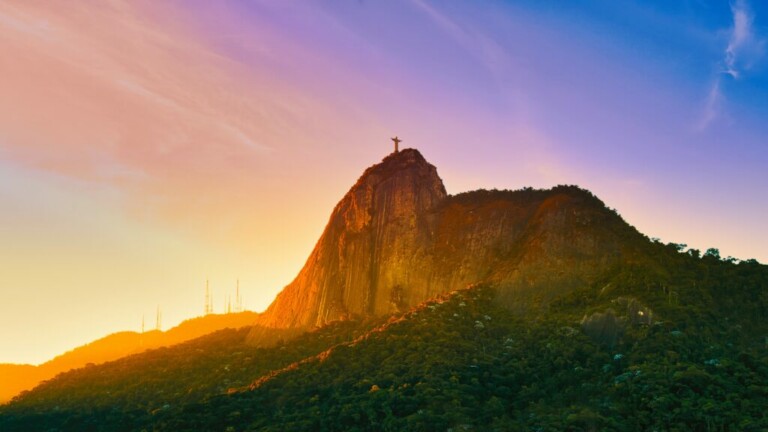 A statue of Christ the Redeemer stands atop Corcovado Mountain in Rio de Janeiro. The mountain, surrounded by a forest and various antennas, overlooks the bustling life of Back Office no Brasil. The sky is a gradient of colors, transitioning from orange to blue.