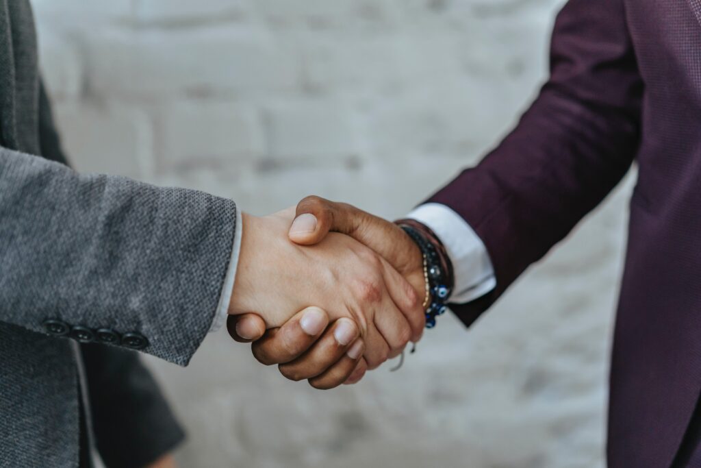 Two individuals are shaking hands against a light-colored brick wall. The person on the left is wearing a gray suit jacket, and the person on the right is wearing a maroon suit jacket and a bracelet. Only their hands and forearms are visible in the image.