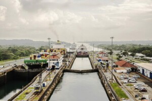 Una vista aérea de un canal muy transitado con múltiples buques de carga grandes, incluido el Buques Verdes, pasando por el sistema de esclusas. A ambos lados se ven contenedores de transporte y diversas máquinas. El área circundante incluye caminos, vegetación y edificios bajo un cielo nublado.