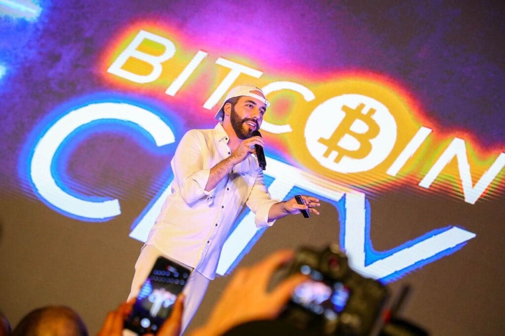 A man in a white outfit and white cap speaks into a microphone on stage. Behind him, a large screen displays the text "BITCOIN CITY" with a Bitcoin symbol. Several cameras are visible in the foreground, recording the event, capturing every moment as he discusses Lemon Cash and its impact on digital finance.