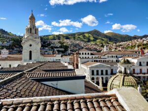 View of a historic city with tiled roofs, a large church with a bell tower, and a dome structure in the foreground. The city, known for having some of the imóveis mais baratos América Latina, is set against a backdrop of hills under a blue sky with scattered clouds.