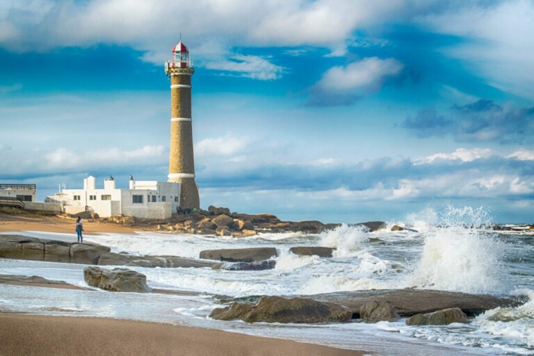 A tall lighthouse stands on a rocky shoreline beside a white building. Waves crash against the rocks, creating splashes. The sky is partly cloudy with patches of blue. A person is seen near the edge of the water, capturing the serene beauty of this EOR no Uruguai scene.