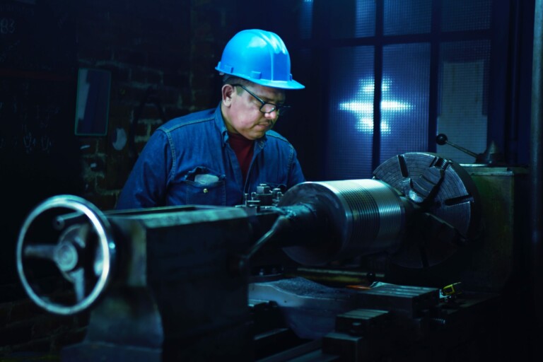 A man wearing a blue hard hat and safety glasses operates a large lathe machine in what appears to be a workshop or industrial setting. With nearshoring in Latin America on the rise, the dimly lit room underscores his focused expression as he diligently attends to his work.