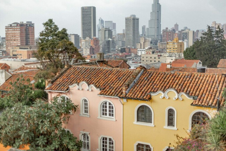 Un paisaje urbano que presenta casas tradicionales con techos de tejas rojas y fachadas coloridas en primer plano, con una mezcla de modernos edificios de gran altura y rascacielos que forman el horizonte de la ciudad al fondo. Se ven árboles a lo largo de las calles, típicos de una zona próspera donde se puede encontrar un abogado en Colombia.