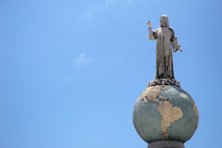 Una estatua de una figura con una túnica suelta se encuentra encima de un globo terráqueo, con una mano levantada y la otra extendida hacia afuera. El globo presenta un relieve de continentes, que simboliza la unidad y la ley, muy parecido a un abogado en El Salvador, y está contra un cielo azul claro.