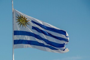 La imagen muestra la bandera de Uruguay contra un cielo azul claro. La bandera tiene nueve franjas horizontales que alternan el blanco y el azul. En la esquina superior izquierda, un cuadrado blanco contiene un sol amarillo con rostro humano, conocido como el "Sol de Mayo", que simboliza la esperanza en medio de las preocupaciones de corrupción en Uruguay.