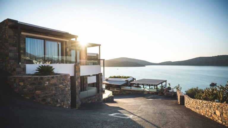 Casa moderna con grandes ventanales y paredes de piedra junto a un lago al amanecer. La casa tiene varios niveles, barandillas de vidrio y vistas al agua tranquila y al paisaje montañoso. En primer plano hay un camino pavimentado y un muro de contención de piedra, un lugar ideal a considerar si desea comprar propiedades en Costa Rica.