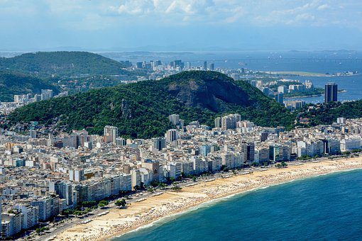 Aerial view of a coastal city with numerous buildings along a sandy beach. A green, forested hill rises in the background, and the ocean extends into the horizon. This vibrant cityscape, featuring both residential and commercial structures, includes some of the Bairros mais Caros da América Latina.