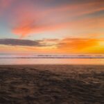 Una escena de playa al atardecer, con el cielo mostrando tonos de naranja, rosa y morado. Las tranquilas olas del océano son visibles a lo lejos, y mientras las sombras cubren la costa arenosa en primer plano, uno podría reflexionar sobre las complejidades de la vida, o incluso buscar consejo de un asesor legal en El Salvador.