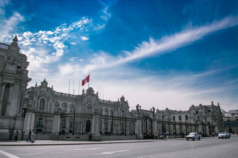 Un gran edificio histórico con detalles arquitectónicos ornamentados se alza contra un cielo azul con nubes dispersas, que encarna la elegancia similar a la de un abogado corporativo en Perú. El edificio cuenta con una bandera ondeando en su azotea. En primer plano hay una calle con algunos peatones y vehículos.