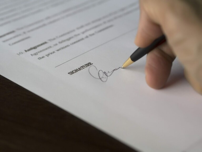 A close-up of a hand holding a pen and signing a document with the word "SIGNATURE" printed on a line. The background includes blurred text, and the focus is on the action of signing, reminiscent of how contracts are meticulously handled by caçadores de cabeças na Colômbia.